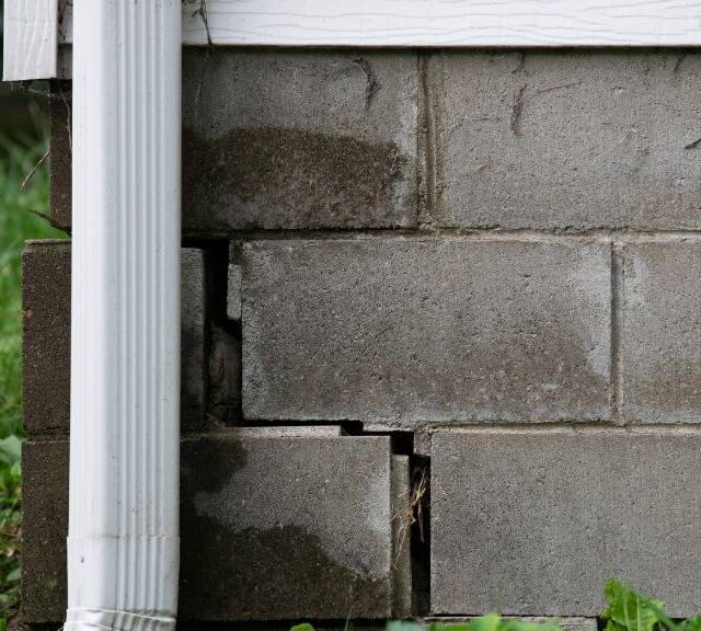 Quality Foundation Repair Austin inspecting stair-step cracks in a concrete block foundation with water damage near a downspo