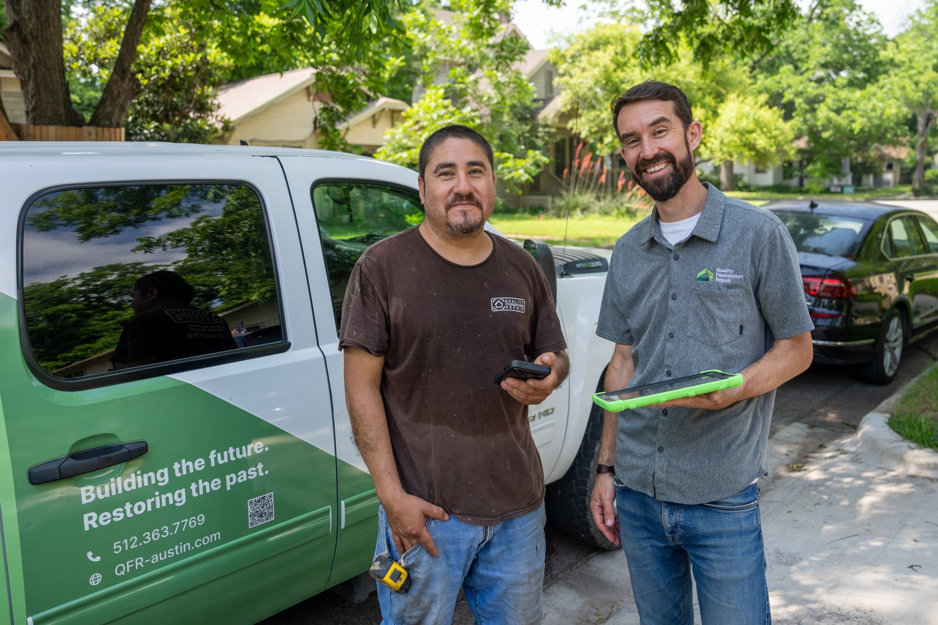Two Quality Foundation Repair Austin team members smiling beside a branded company truck, one holding a tablet.
